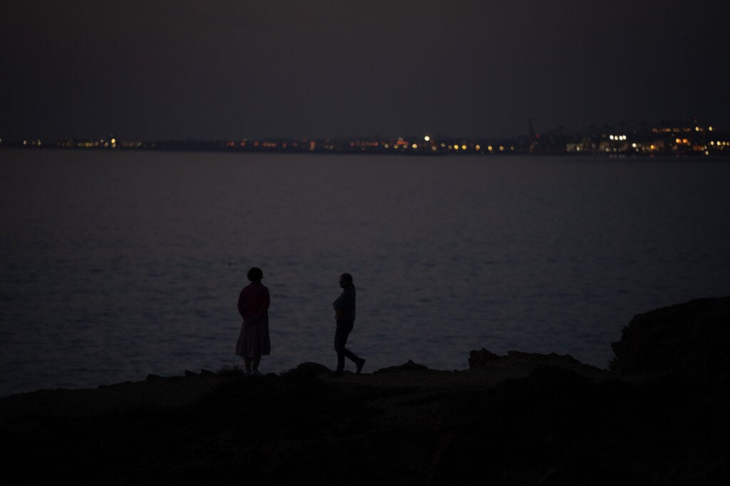 Silhouettes of two people standing on rocks by the water at night with city lights in the background.