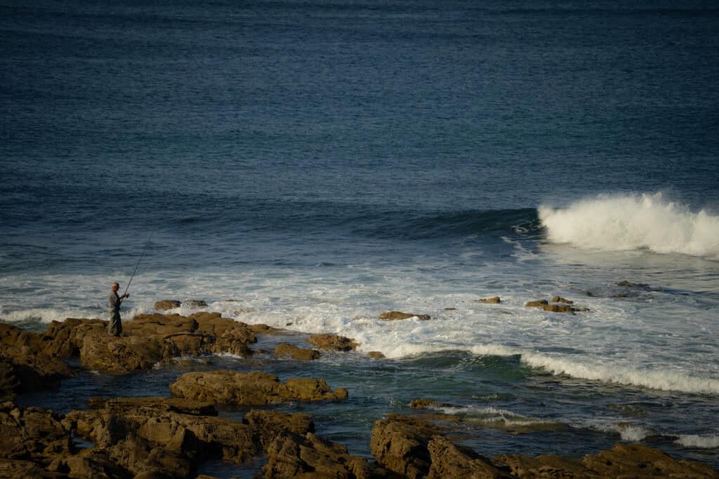 Fisherman standing on rocks fishing by the ocean at sunset.